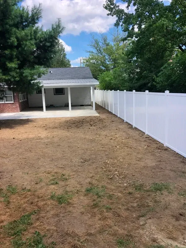 Backyard with dirt yard, white fence, trees, and a small covered patio shed under a partly cloudy sky