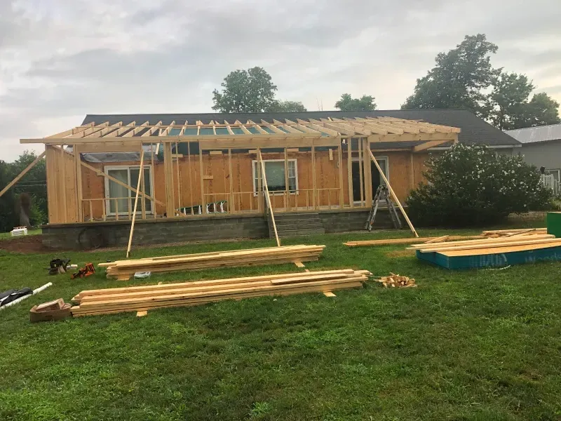 House under construction with wooden framing, roof trusses, scaffolding, and lumber stacked on the grass