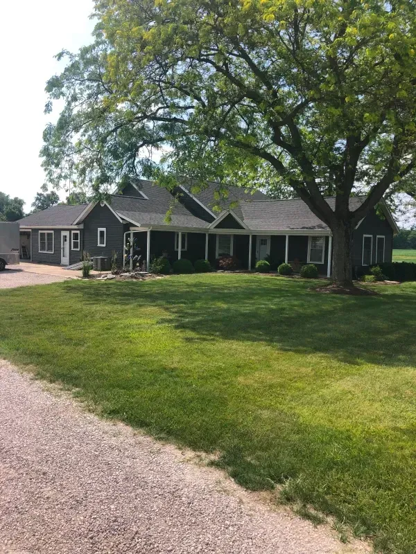 Single-story dark house with a large front lawn and a tree beside a gravel driveway