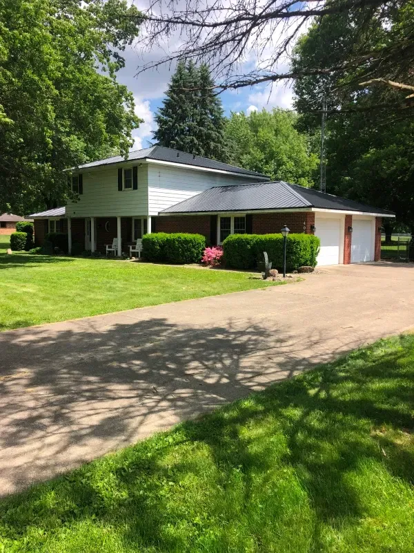 Two-story suburban house with white siding, red trim, attached garage, and a tree-lined driveway