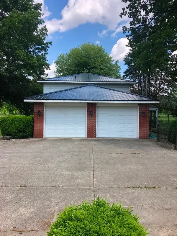Two-car garage with white doors, red brick pillars, and a metal roof, framed by trees and a concrete driveway.
