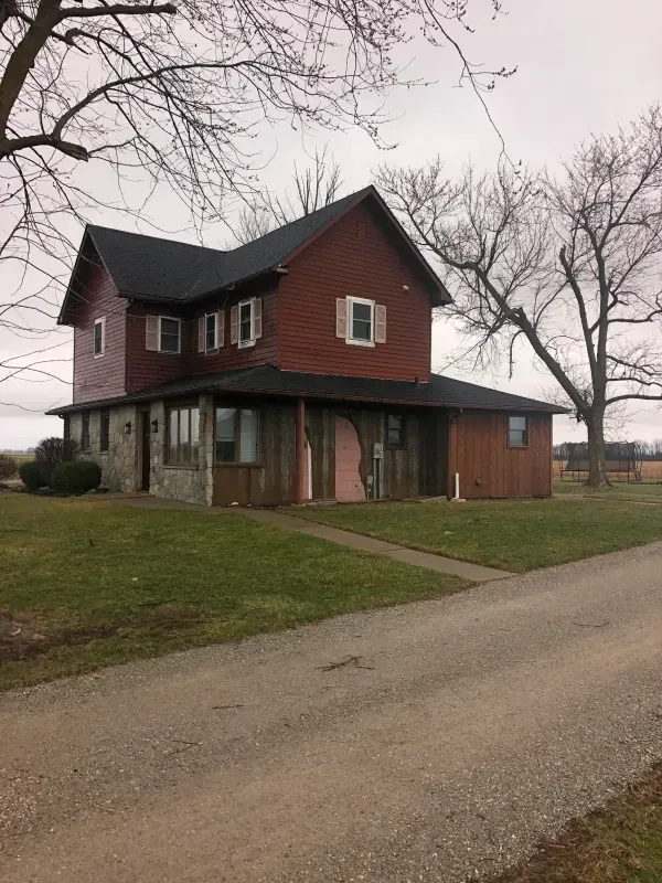 Two-story red house with a covered porch beside a gravel road on an overcast day