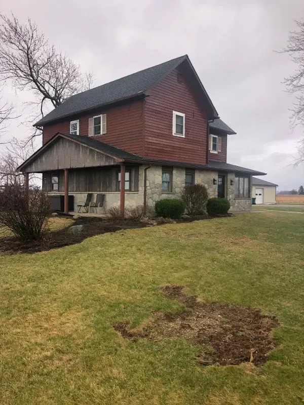 Two-story farmhouse with red siding and porch on a grassy lot under a cloudy sky
