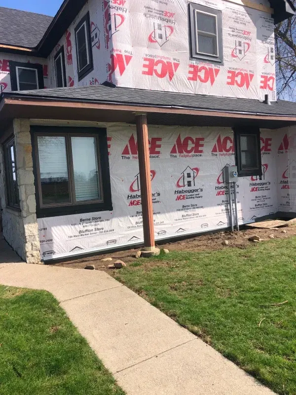 House under construction wrapped in white housewrap, with a front window, porch post, and sidewalk in front