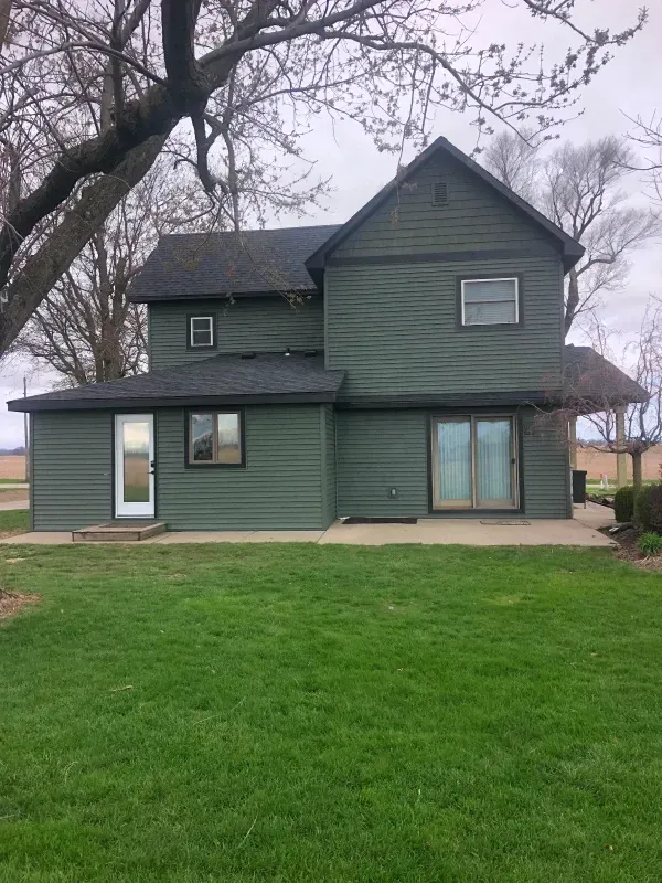 Green two-story house with a small porch and large front window on a grassy lot under overcast sky