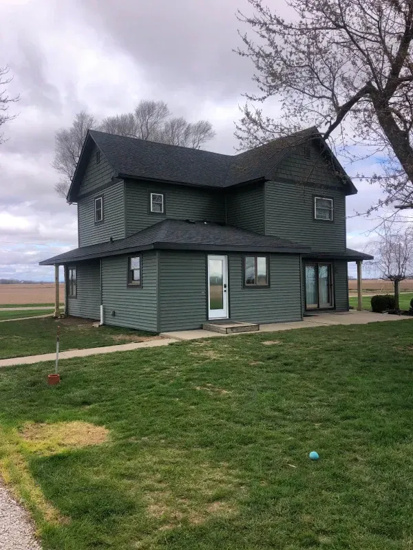 Dark green two-story farmhouse with a porch in a grassy field under a cloudy sky