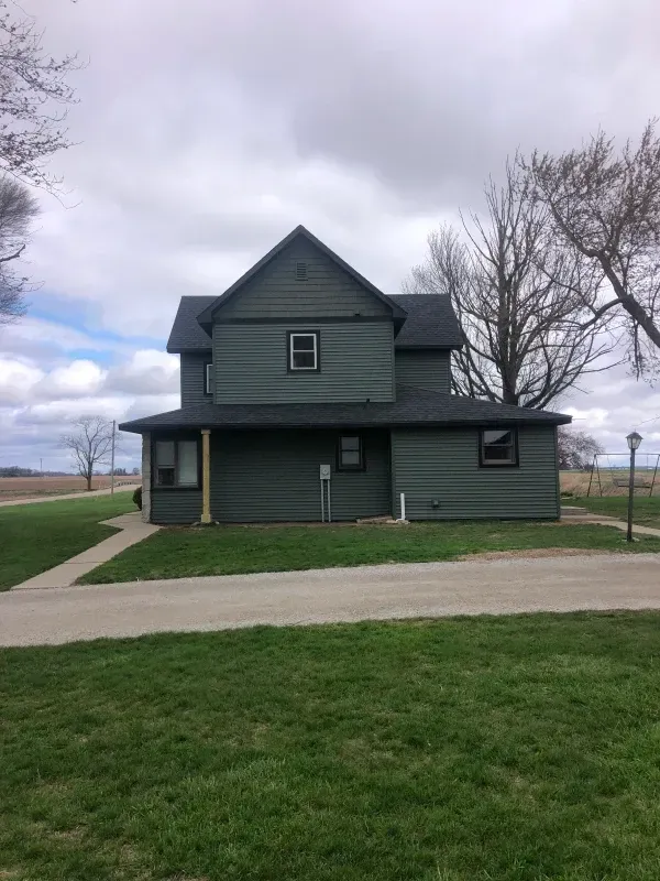 Dark green two-story farmhouse with porch, set beside a road under a cloudy sky.
