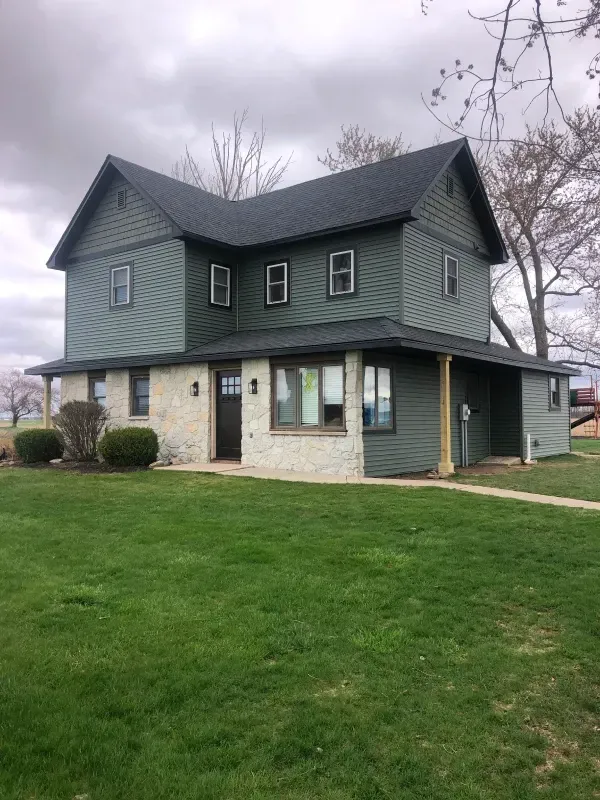 Two-story house with green siding, stone accents, and a large grassy lawn under an overcast sky