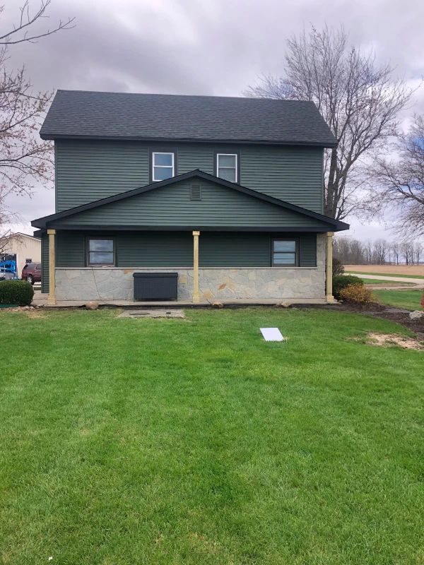 Two-story green house with dark roof, front porch, and lawn under an overcast sky