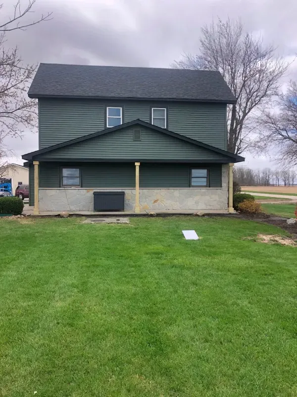 Dark green house with a covered front porch and a large lawn under an overcast sky