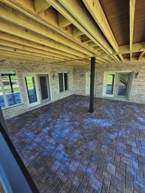 Covered brick patio under a wooden deck, with stone walls, windows, and a central support post.
