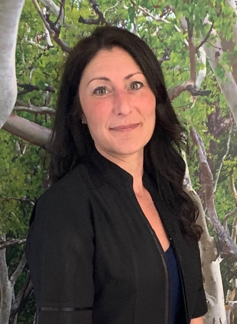 A woman in a black jacket is standing in front of a painting of trees.