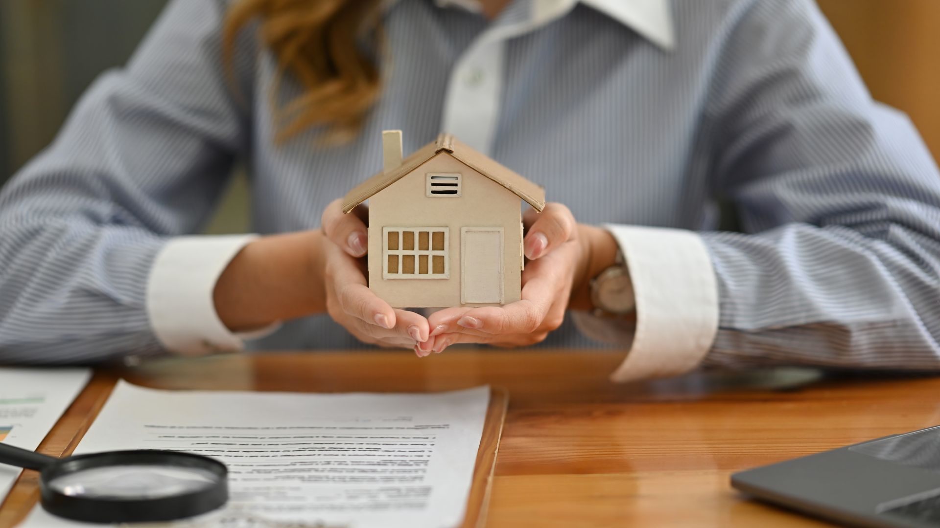 Person holding a miniature house above documents and a laptop, suggesting a real estate transaction.