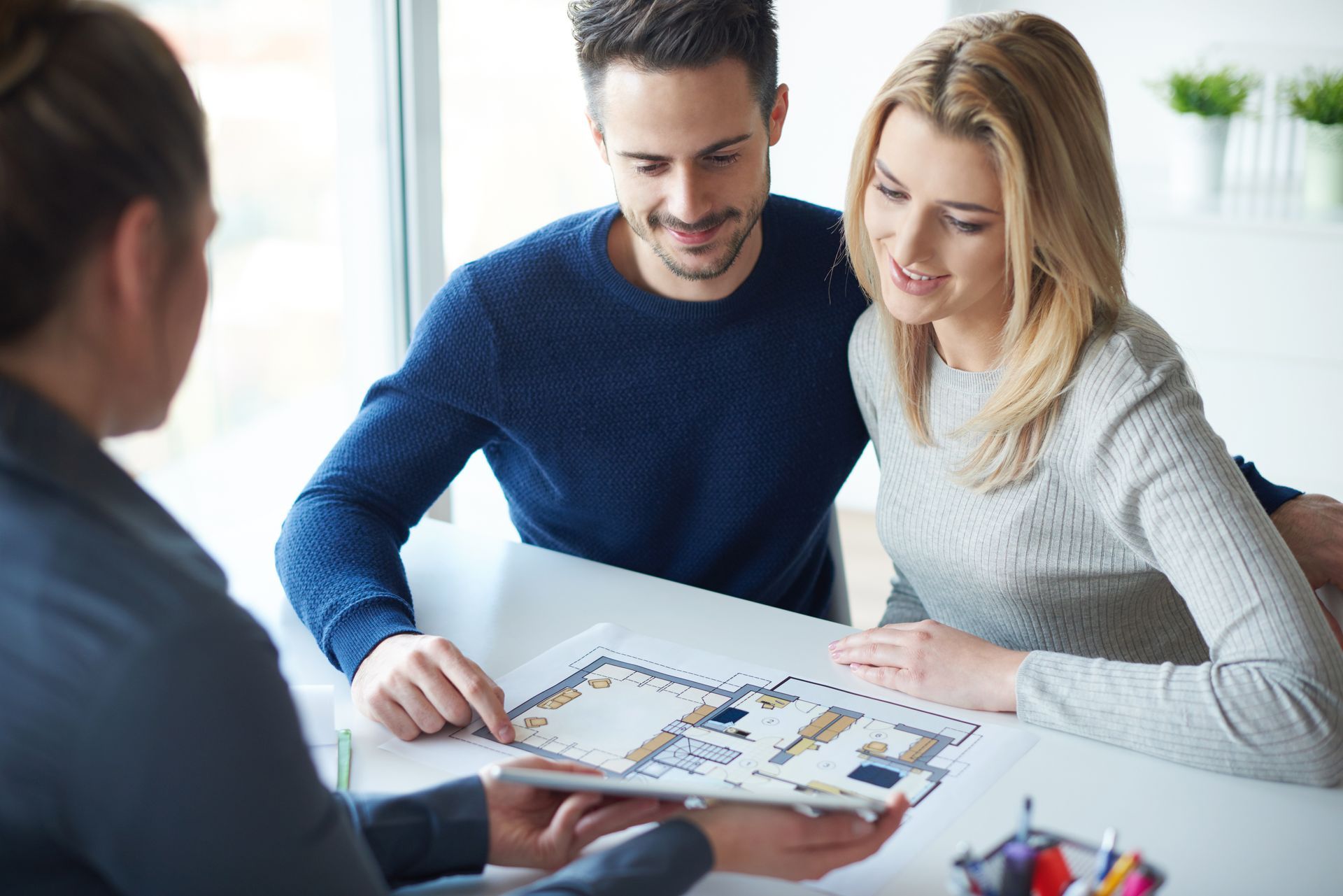 Couple reviews a floor plan with a professional at a table. The man points; they smile. Couple reviews a floor plan with a professional at a table. The man points; they smile.