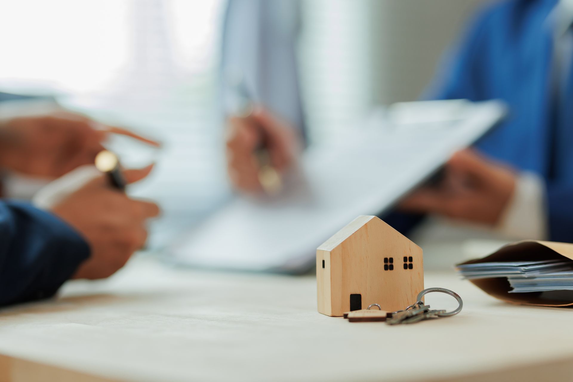 Wooden house model with keys on a table, with people in business attire signing documents in the background.