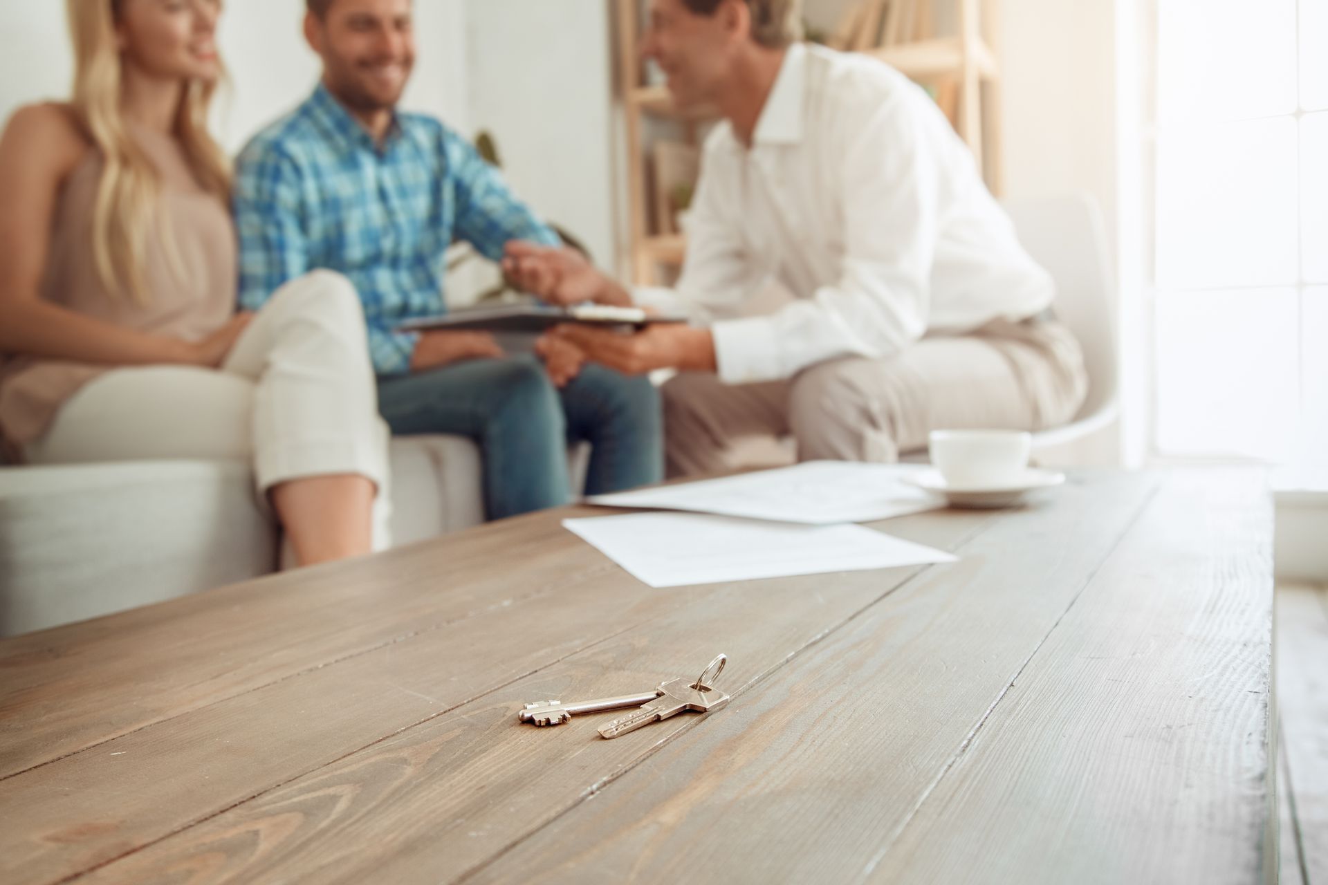 Keys on a wooden table with a couple and a man in the background, possibly discussing a property deal. Keys on a wooden table with a couple and a man in the background, possibly discussing a property deal.