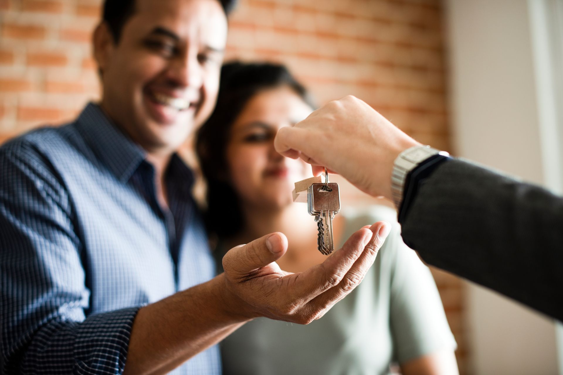 Couple receiving keys from a person, likely a real estate agent, with a brick wall background. Couple receiving keys from a person, likely a real estate agent, with a brick wall background.