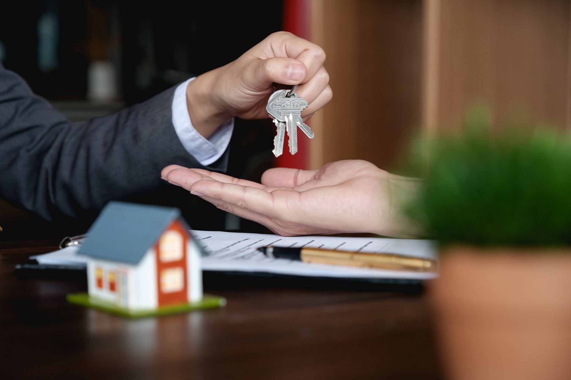 Person handing keys to another, symbolizing a real estate transaction. A house model and paperwork are on the table.