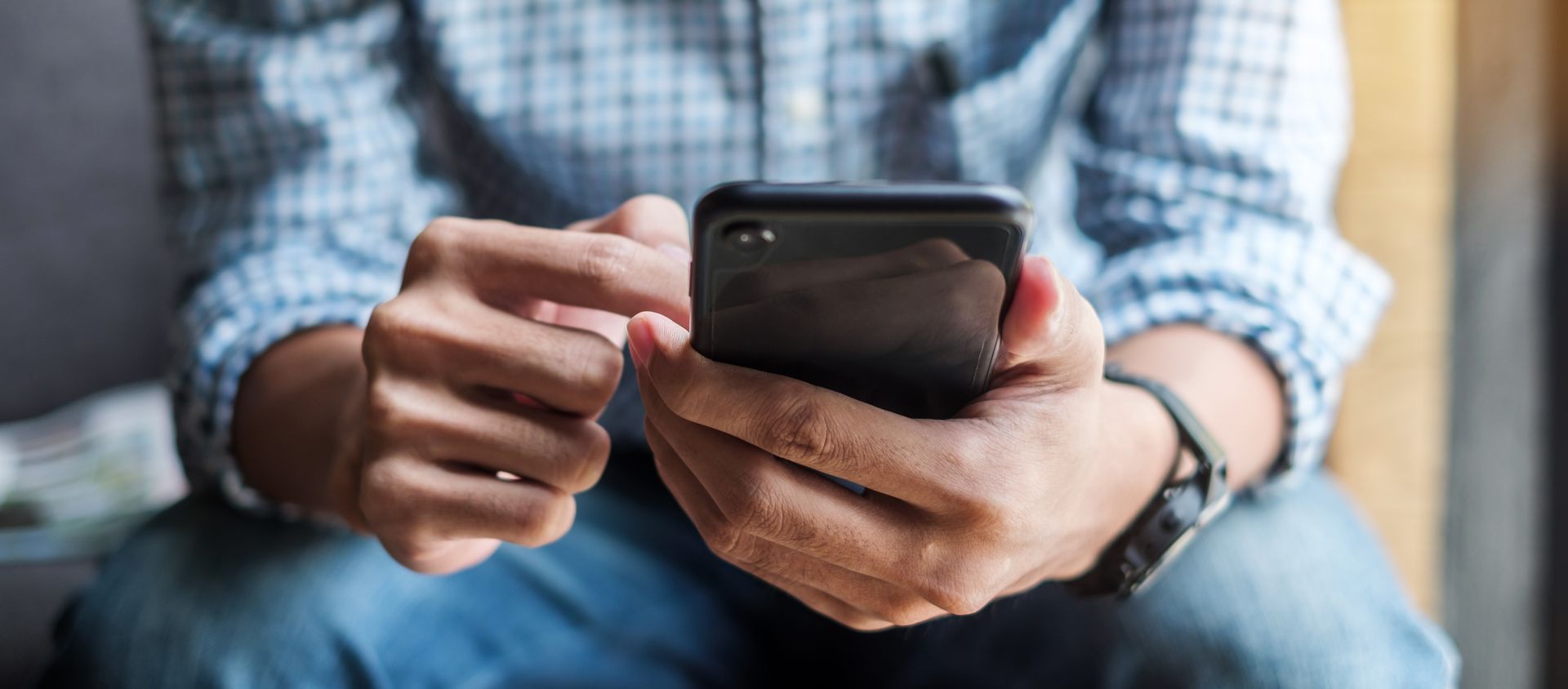 A person using a black smartphone while sitting down. They are wearing a blue checkered shirt and a watch.