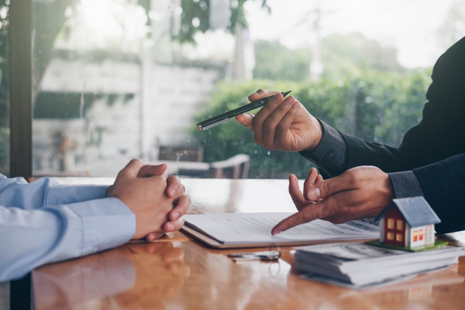 Hands pointing at a contract and a small house model. Two people are seated at a table. Hands pointing at a contract and a small house model. Two people are seated at a table.