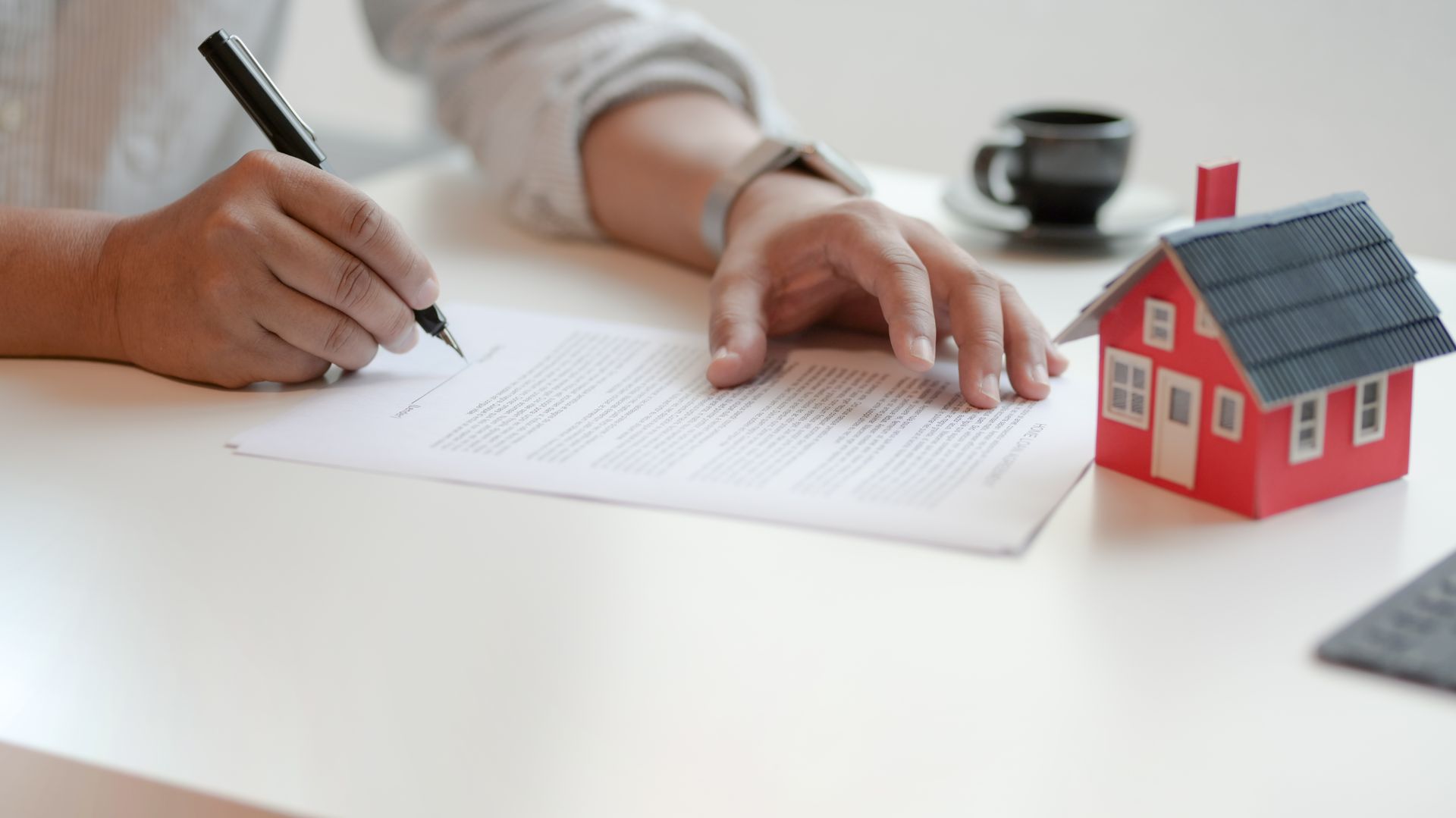 Person signs document on a white desk next to a model house and coffee cup. Person signs document on a white desk next to a model house and coffee cup.