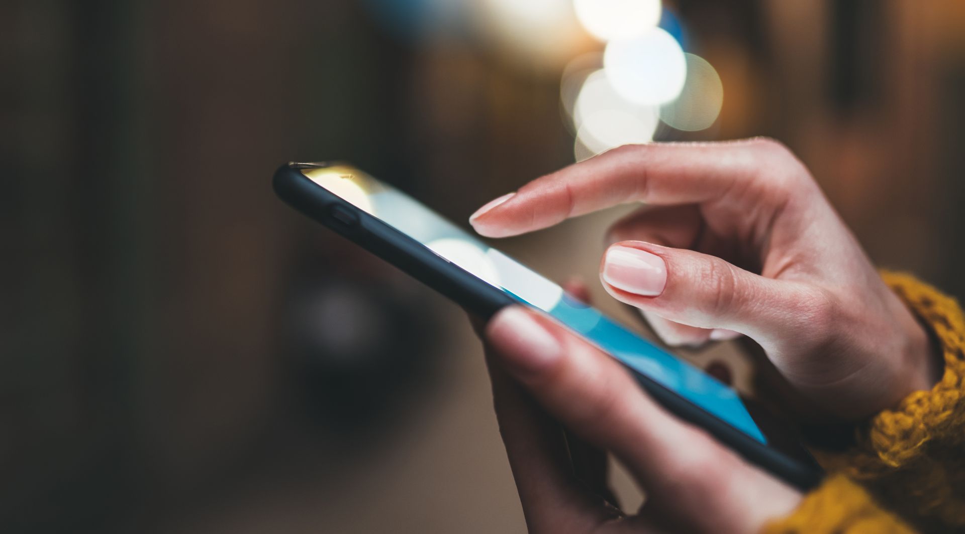 Person's hands using a smartphone outdoors at night. Bright screen illuminates fingers as they scroll.