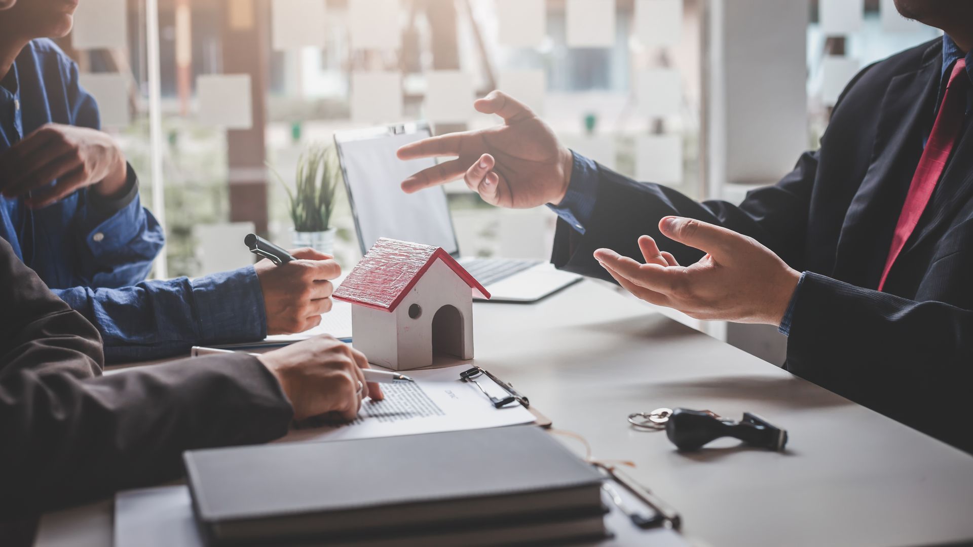 People discussing paperwork around a table with a house model, keys, and laptop.