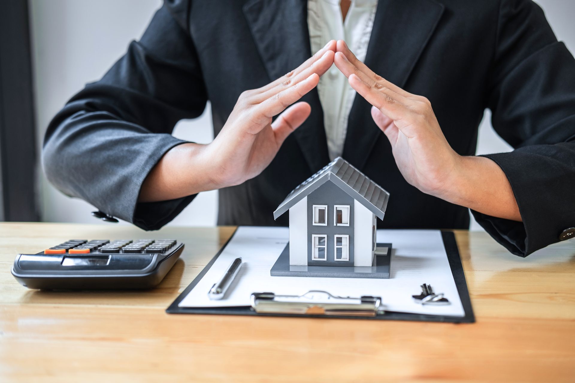 Hands sheltering a miniature house on a clipboard with a calculator and pen, symbolizing protection or insurance.