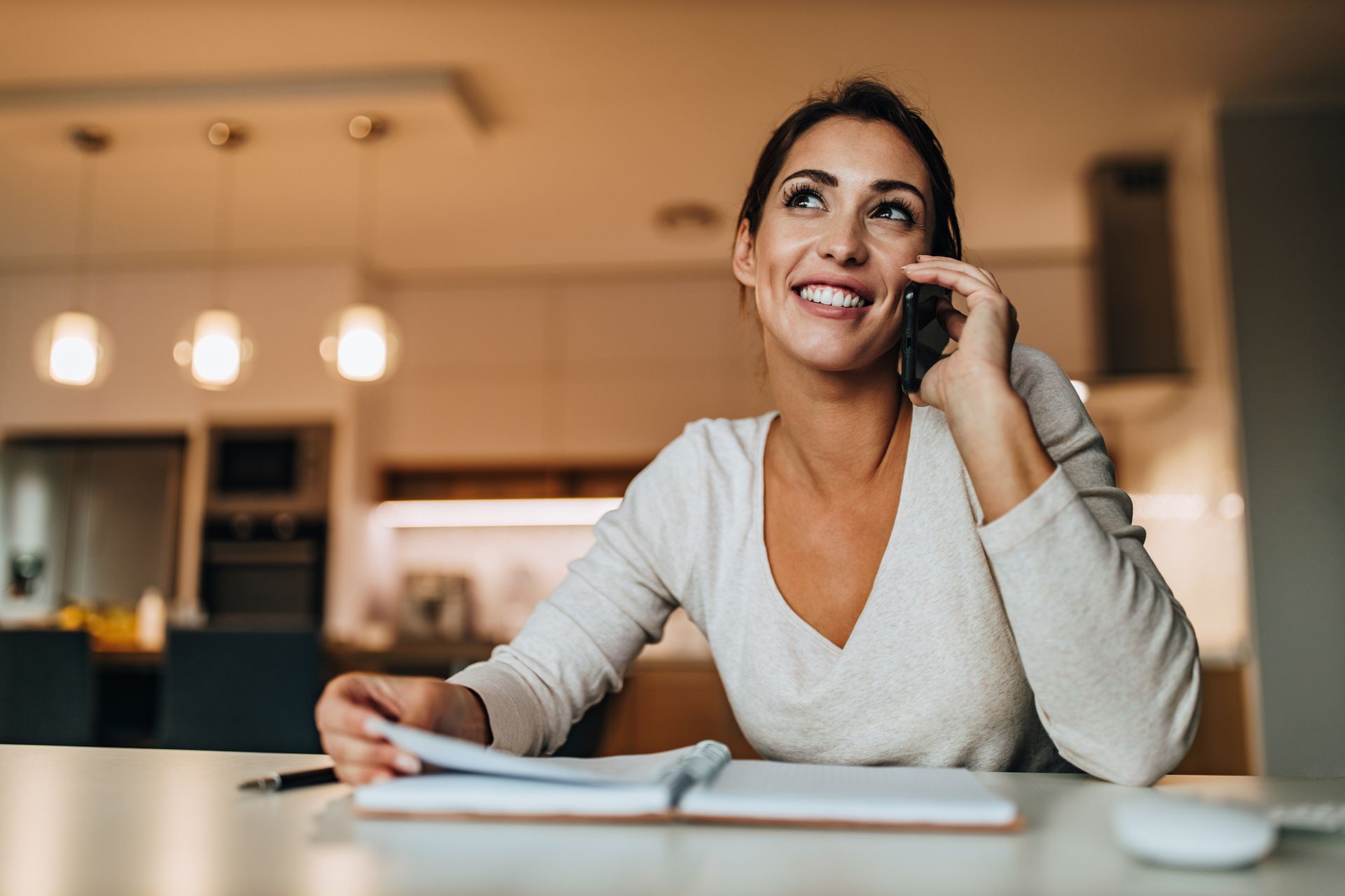 Woman smiling while talking on a phone, looking upward, sitting at a table with a notebook in a modern kitchen.