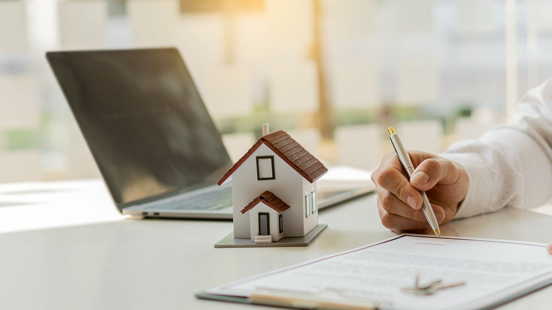 Person signing a document, with a laptop, model house, and keys on the table. Person signing a document, with a laptop, model house, and keys on the table.