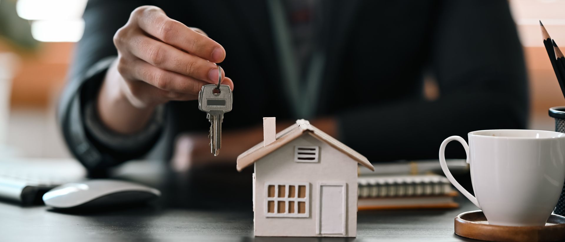 Hand holding keys over a model house; real estate concept. Desk with a cup, pen, and mouse in the background. Hand holding keys over a model house; real estate concept. Desk with a cup, pen, and mouse in the background.