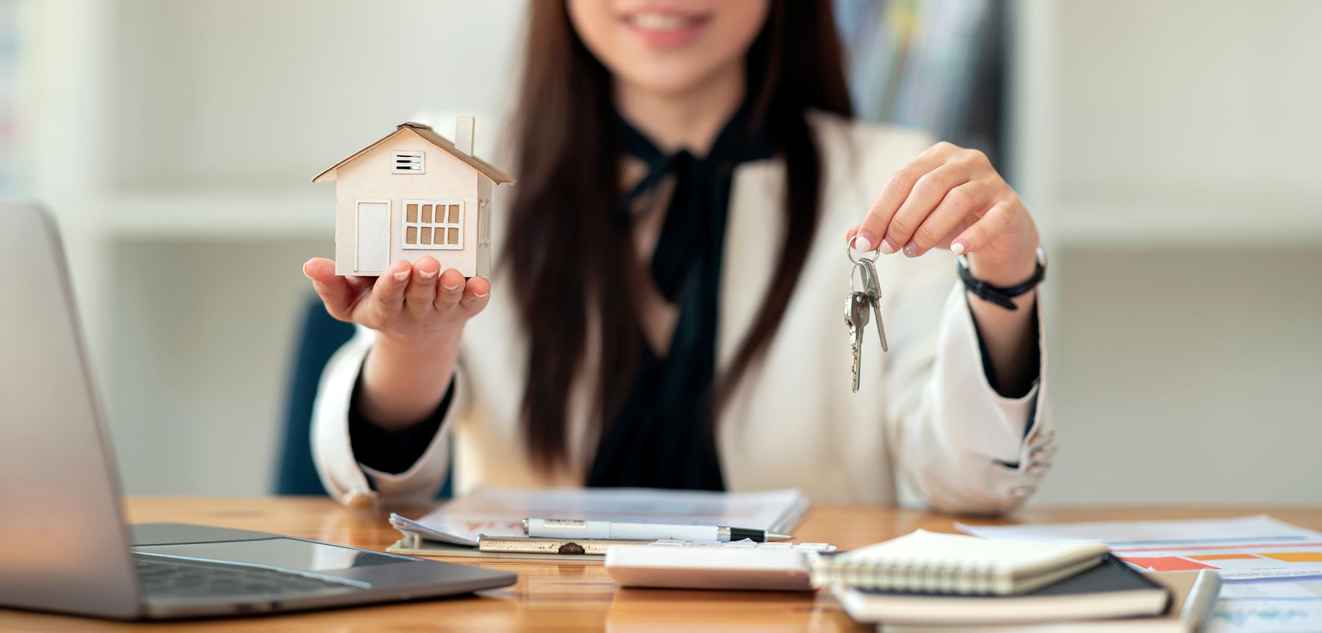 A person holding a miniature house and keys, smiling at a desk with a laptop and documents.