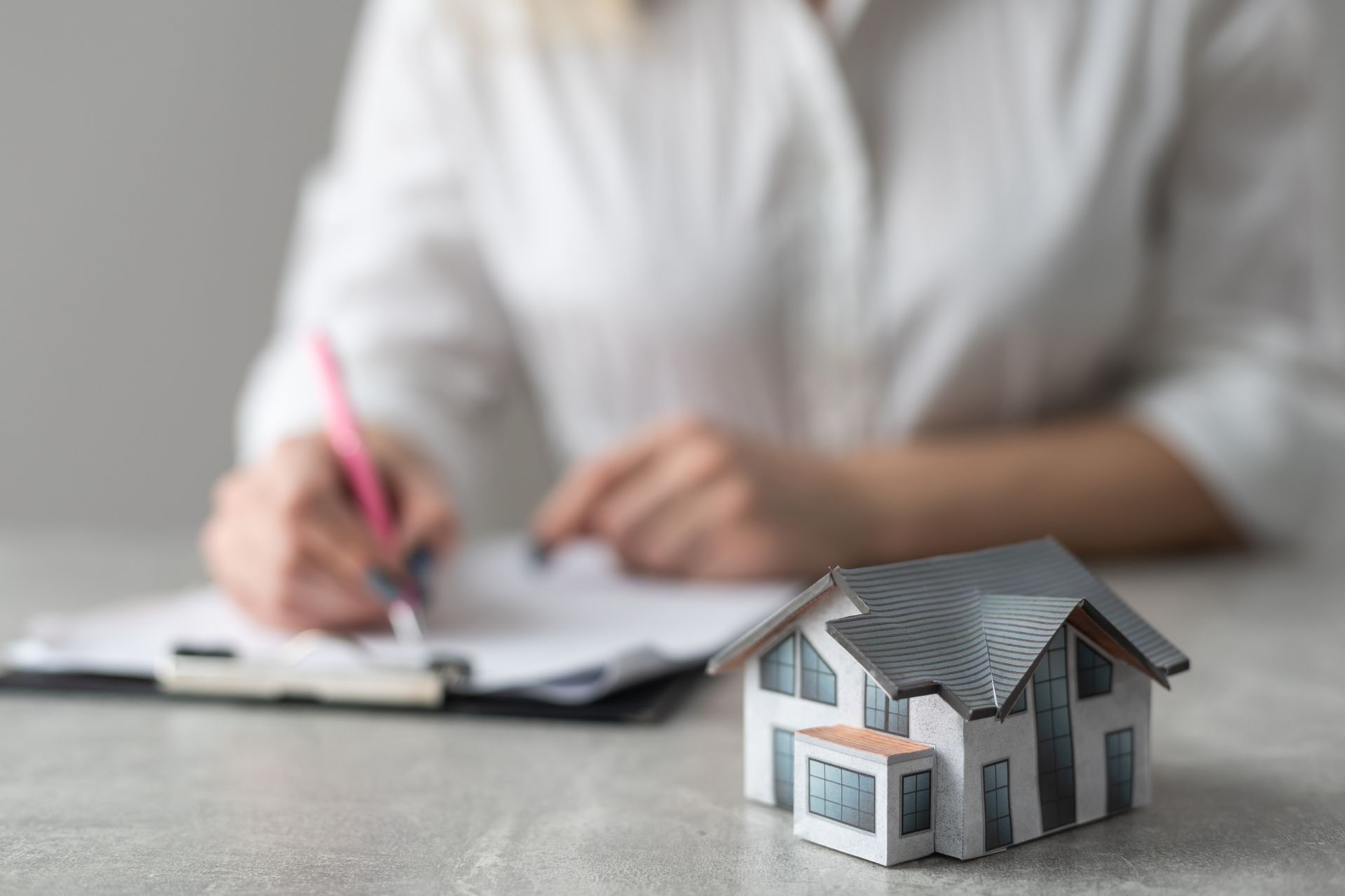 Miniature house in front of person writing on paper, possibly a real estate transaction. Miniature house in front of person writing on paper, possibly a real estate transaction.