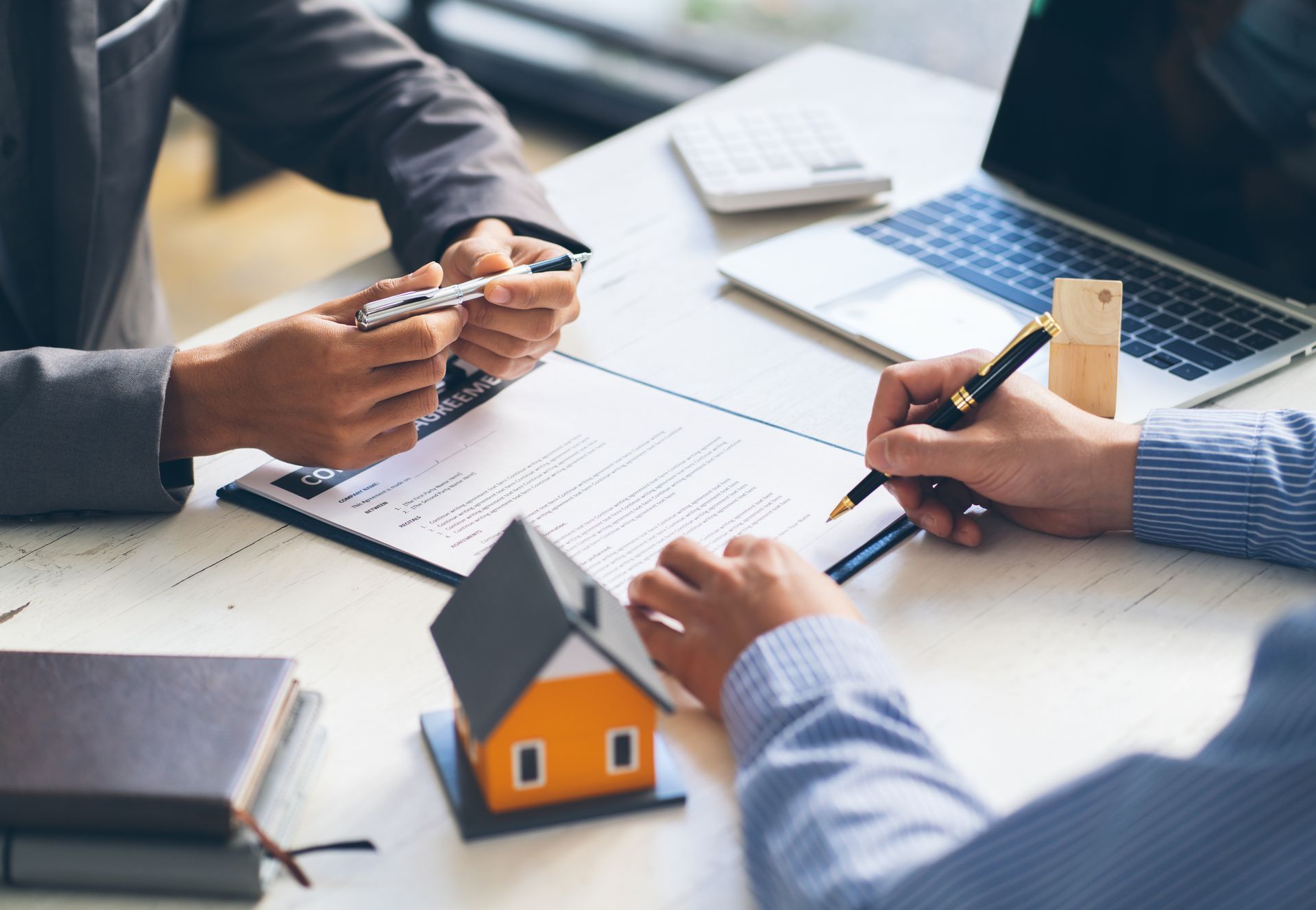 Person signing a document, home model on desk, laptop, calculator, business setting.
