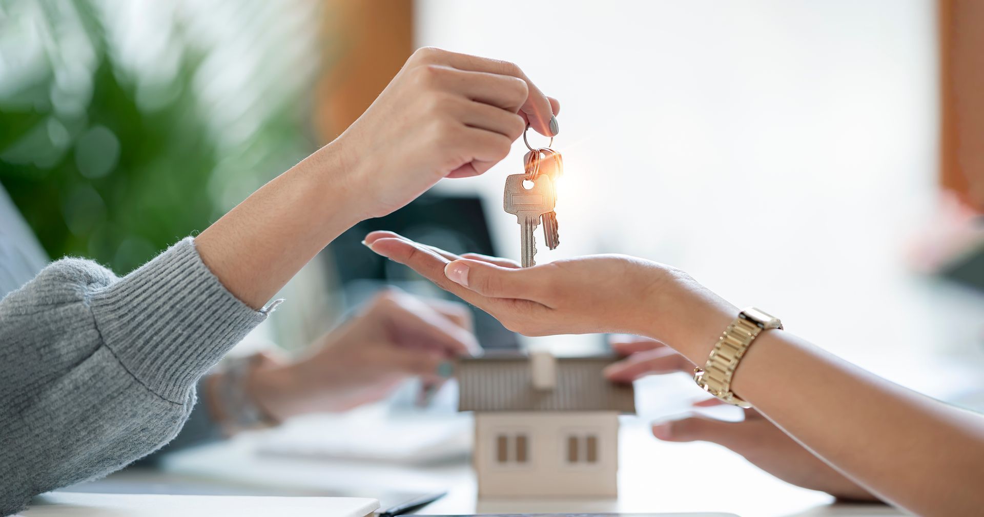 Person handing keys over a miniature house model, symbolizing a real estate transaction.