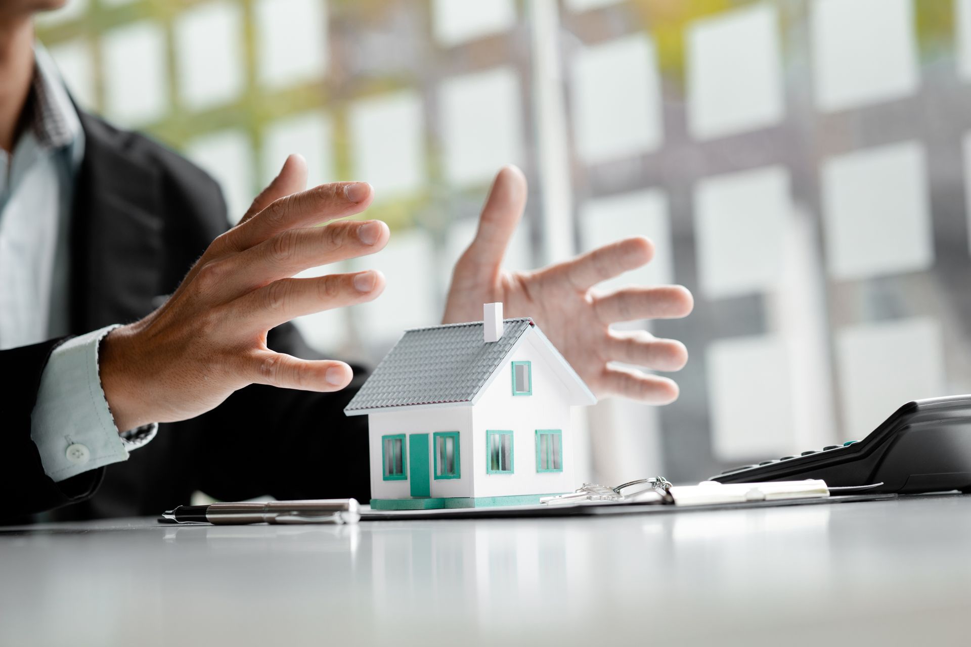 Person gesturing towards a model house on a desk with a calculator.