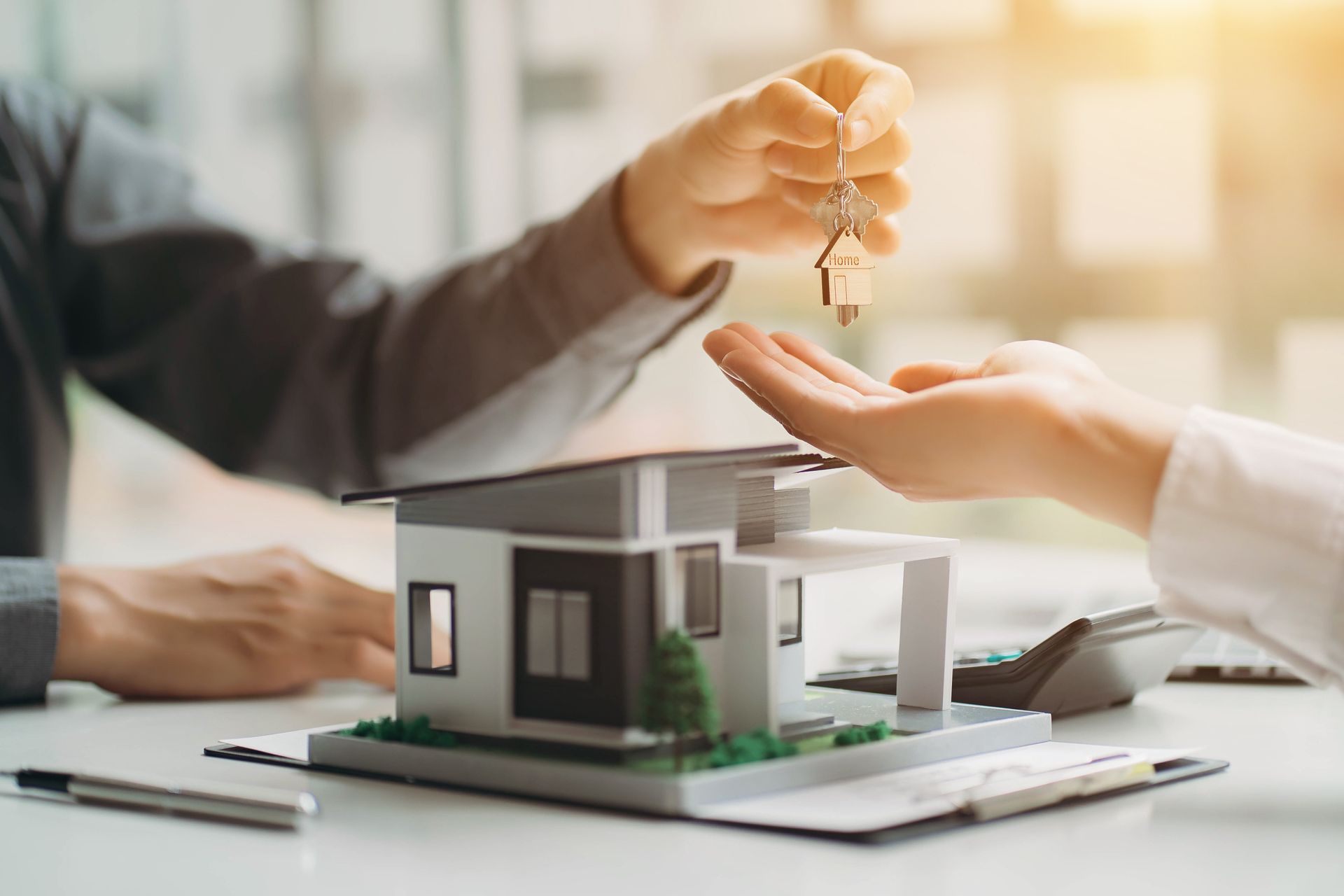 Person handing over house keys to another person over a model house; sunlight.