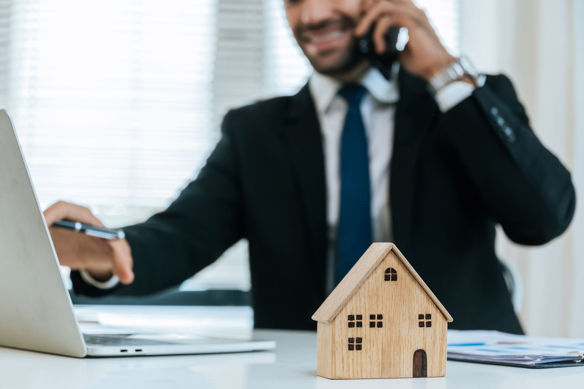 Real estate agent on phone, working on laptop with small wooden house on desk. Real estate agent on phone, working on laptop with small wooden house on desk.