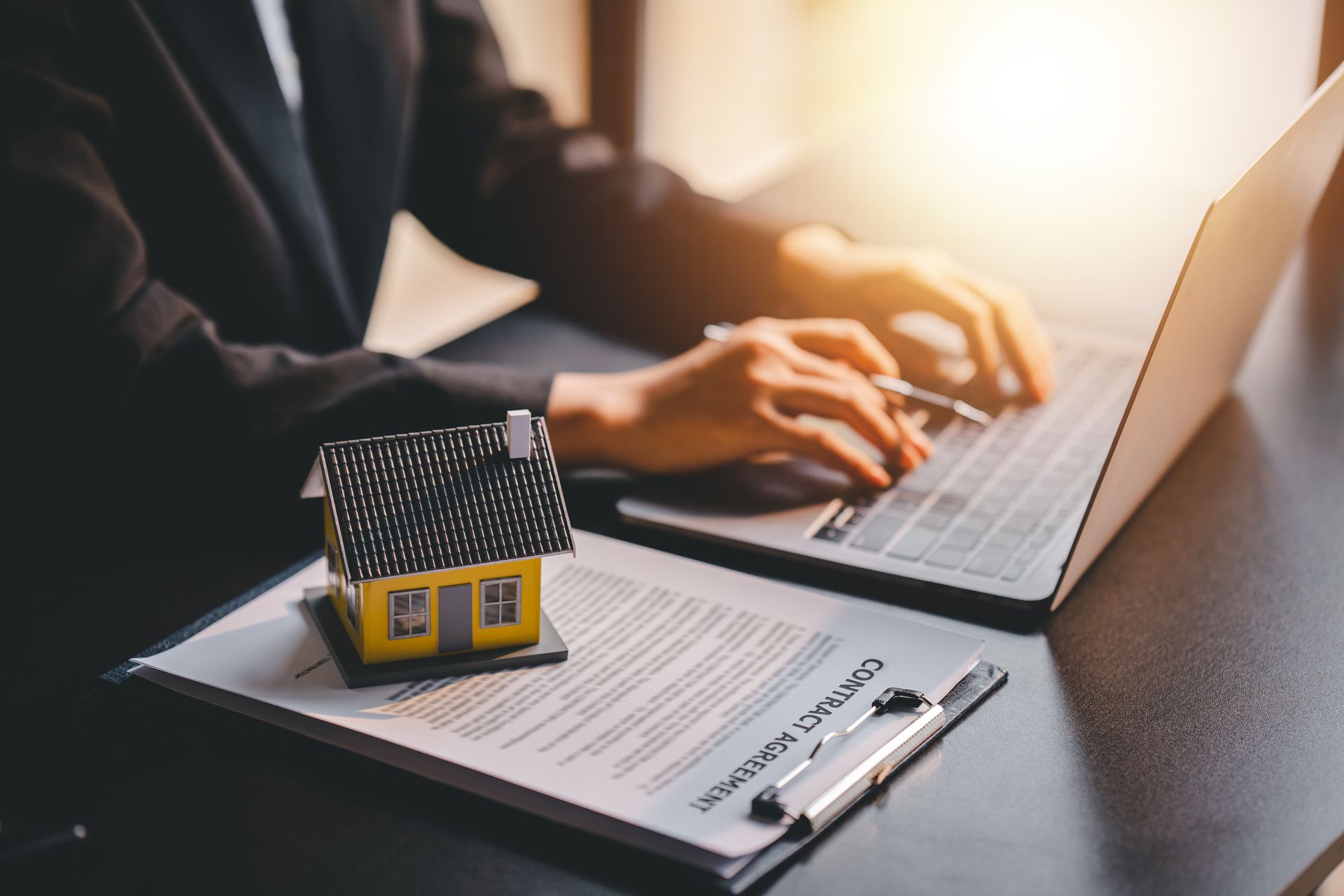 Person typing on laptop, house model and contract on desk, sunlight.