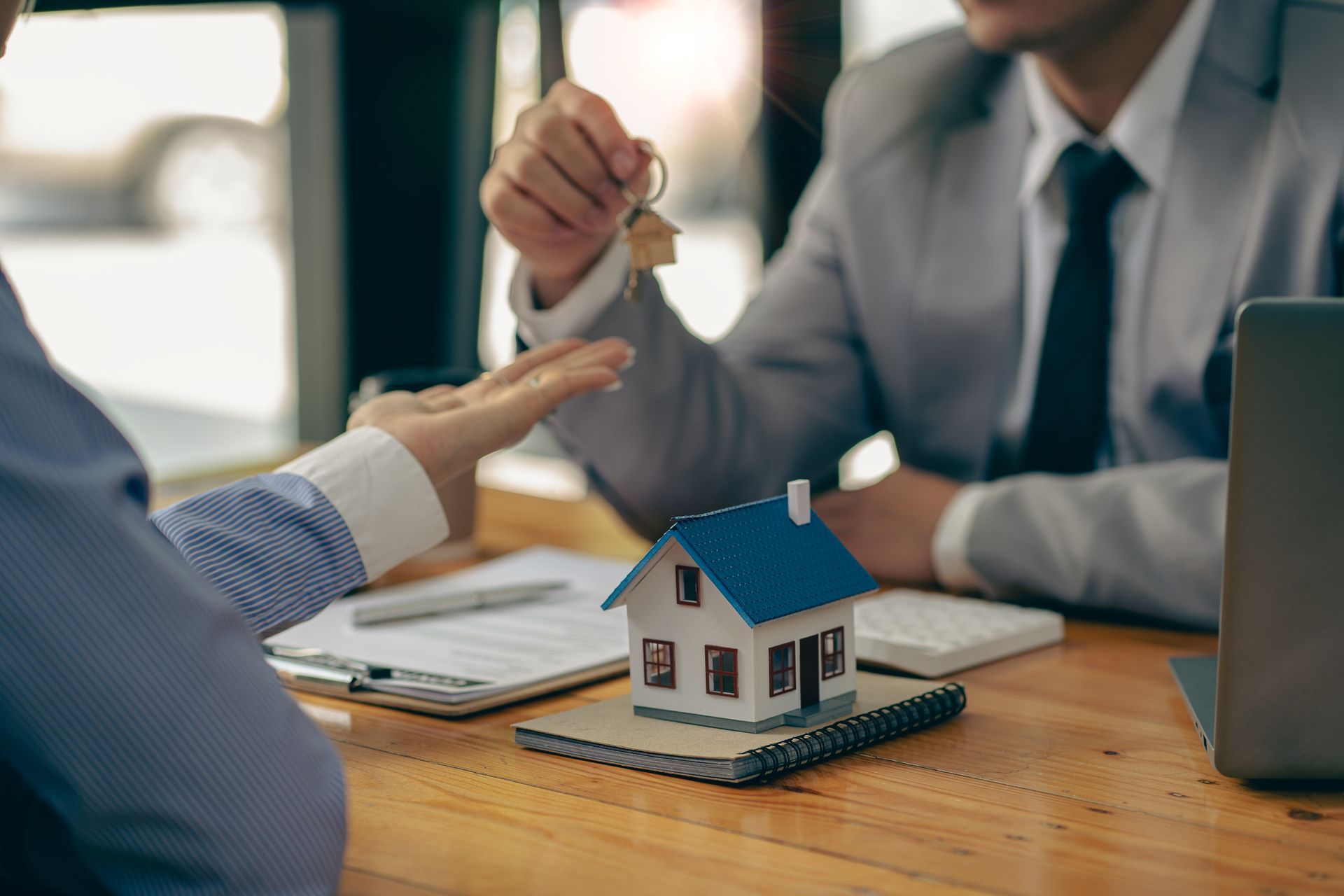 Real estate agent handing keys to a person, over a small house model and paperwork on a desk.