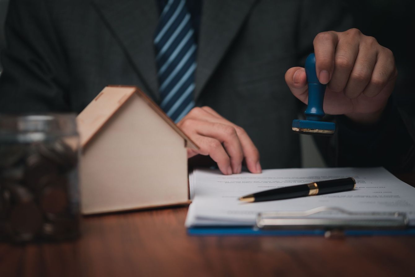 Person stamping a document with a blue stamp; small house model and jar of coins on the table. Person stamping a document with a blue stamp; small house model and jar of coins on the table.