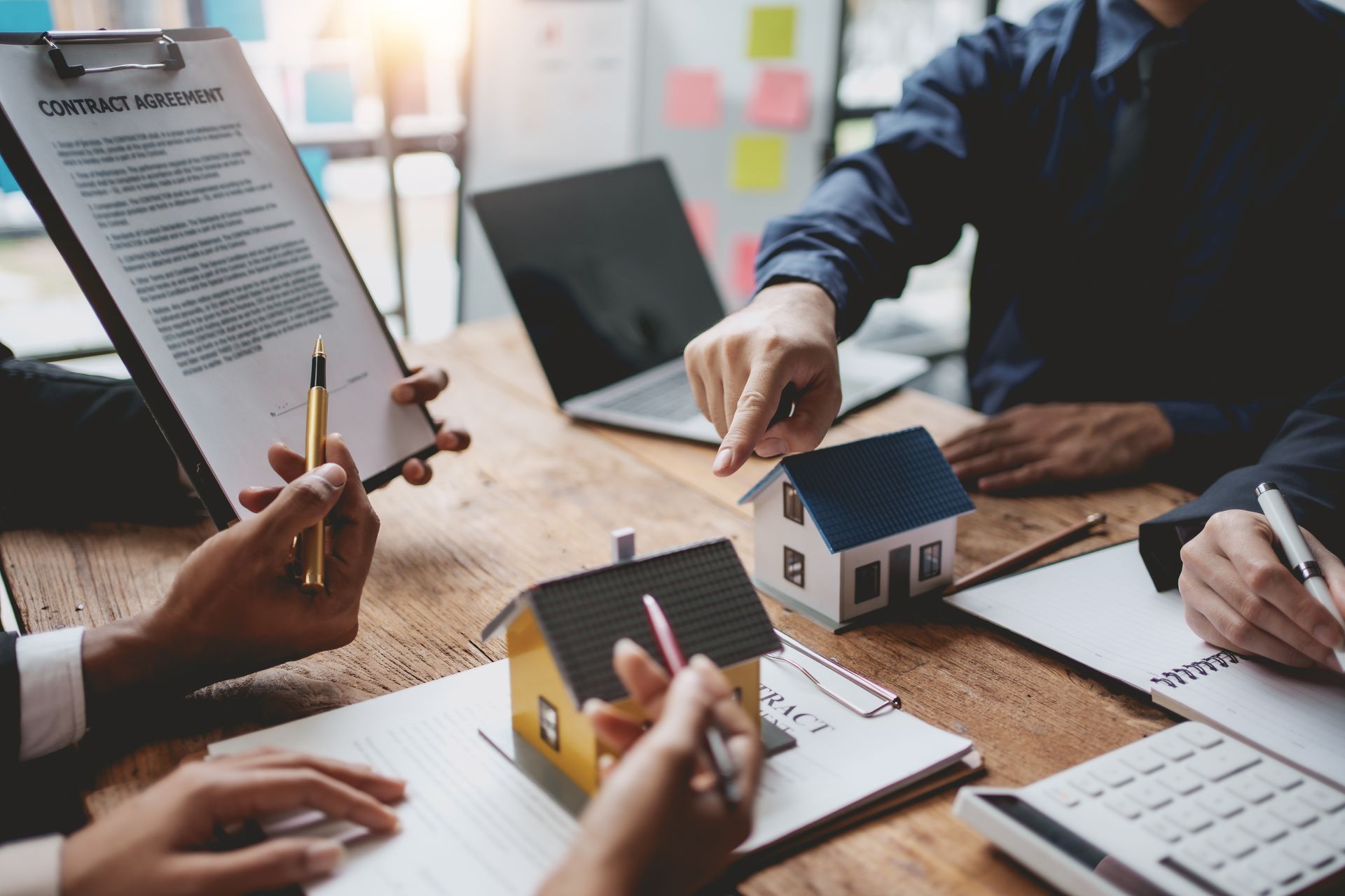People reviewing documents, pointing at toy houses, and using a laptop at a table, likely discussing a real estate deal. People reviewing documents, pointing at toy houses, and using a laptop at a table, likely discussing a real estate deal.