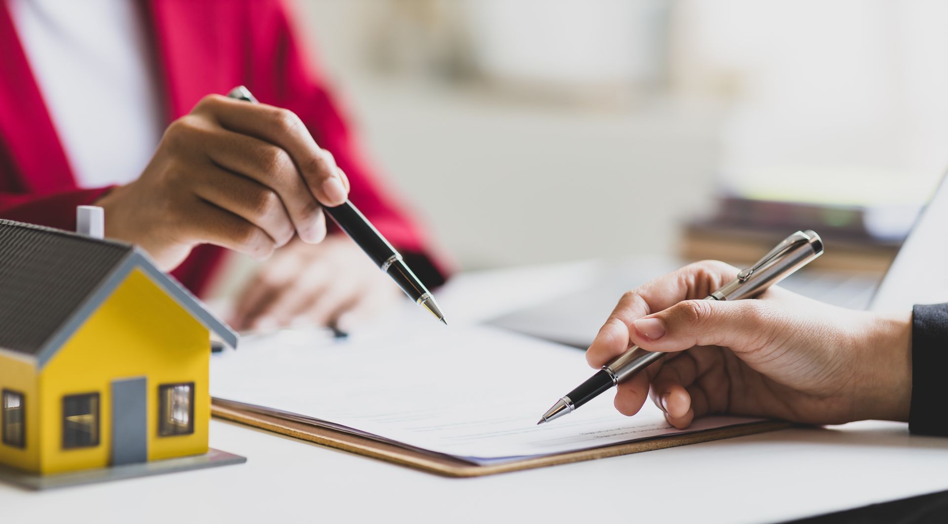 Hands signing a document, miniature yellow house, pen pointing.