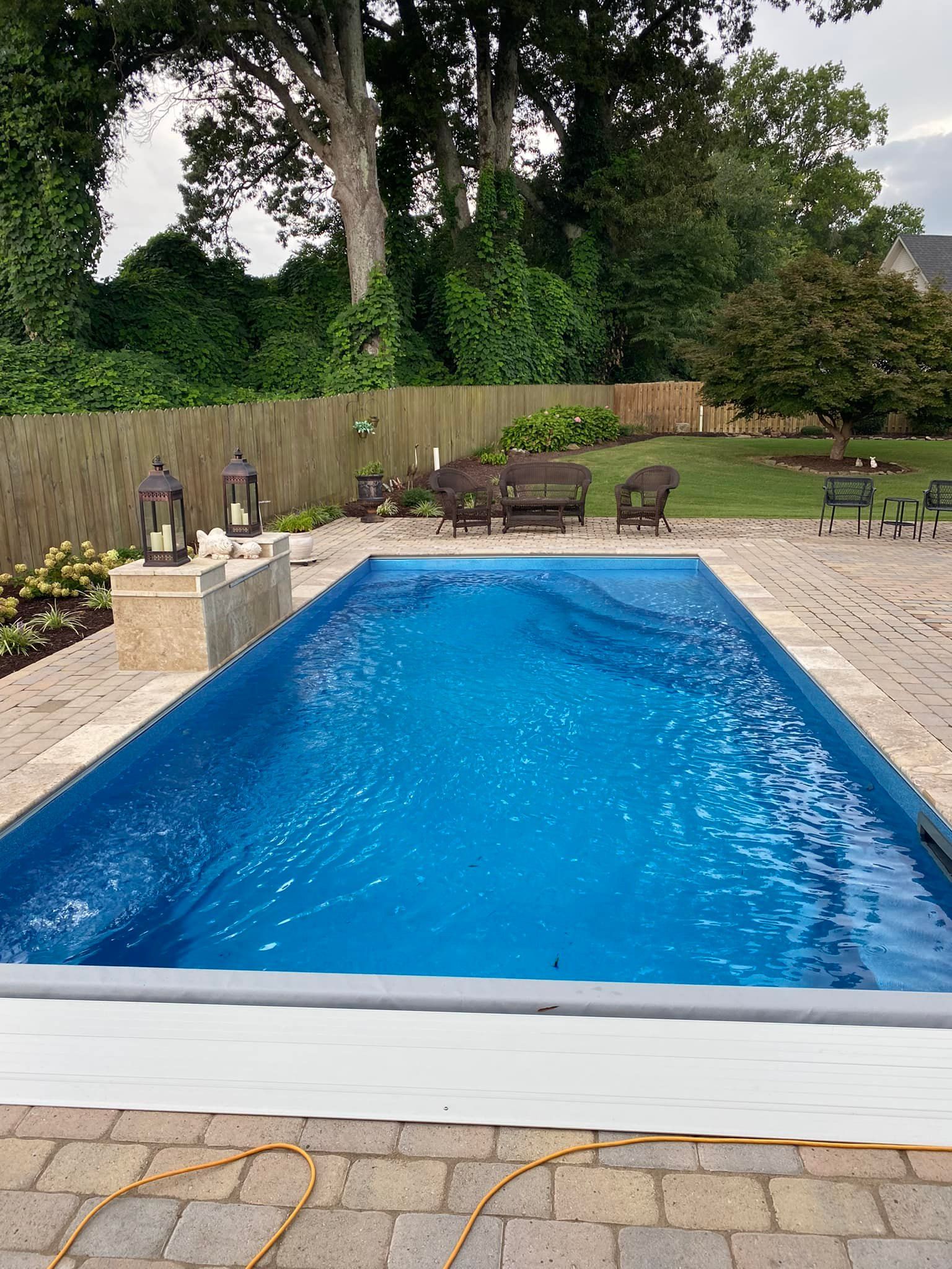 Rectangular swimming pool surrounded by brick patio, with seating area in the background and foliage.