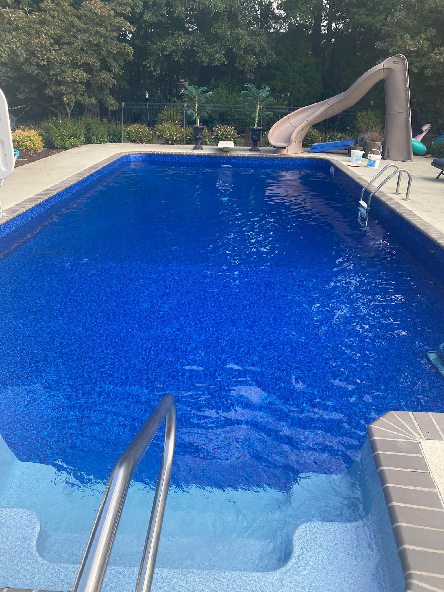 Blue-tiled rectangular pool with a slide, surrounded by a concrete patio and trees.