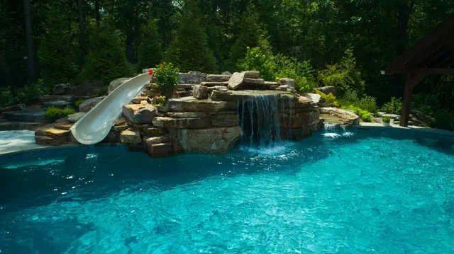Swimming pool with waterfall and slide, surrounded by rocks and greenery.