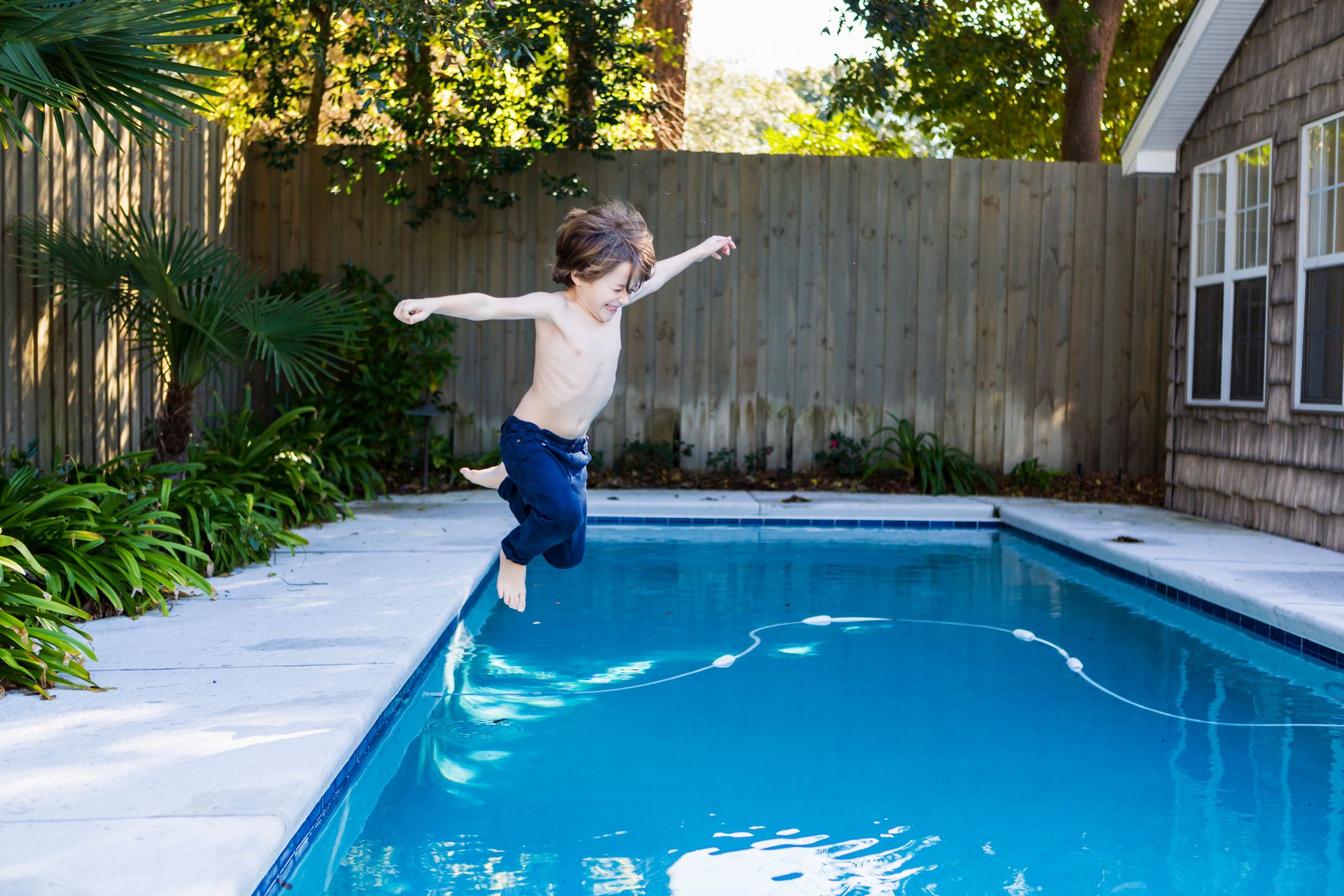Boy jumps excitedly into a pool, arms outstretched. Blue water, sunny backyard.