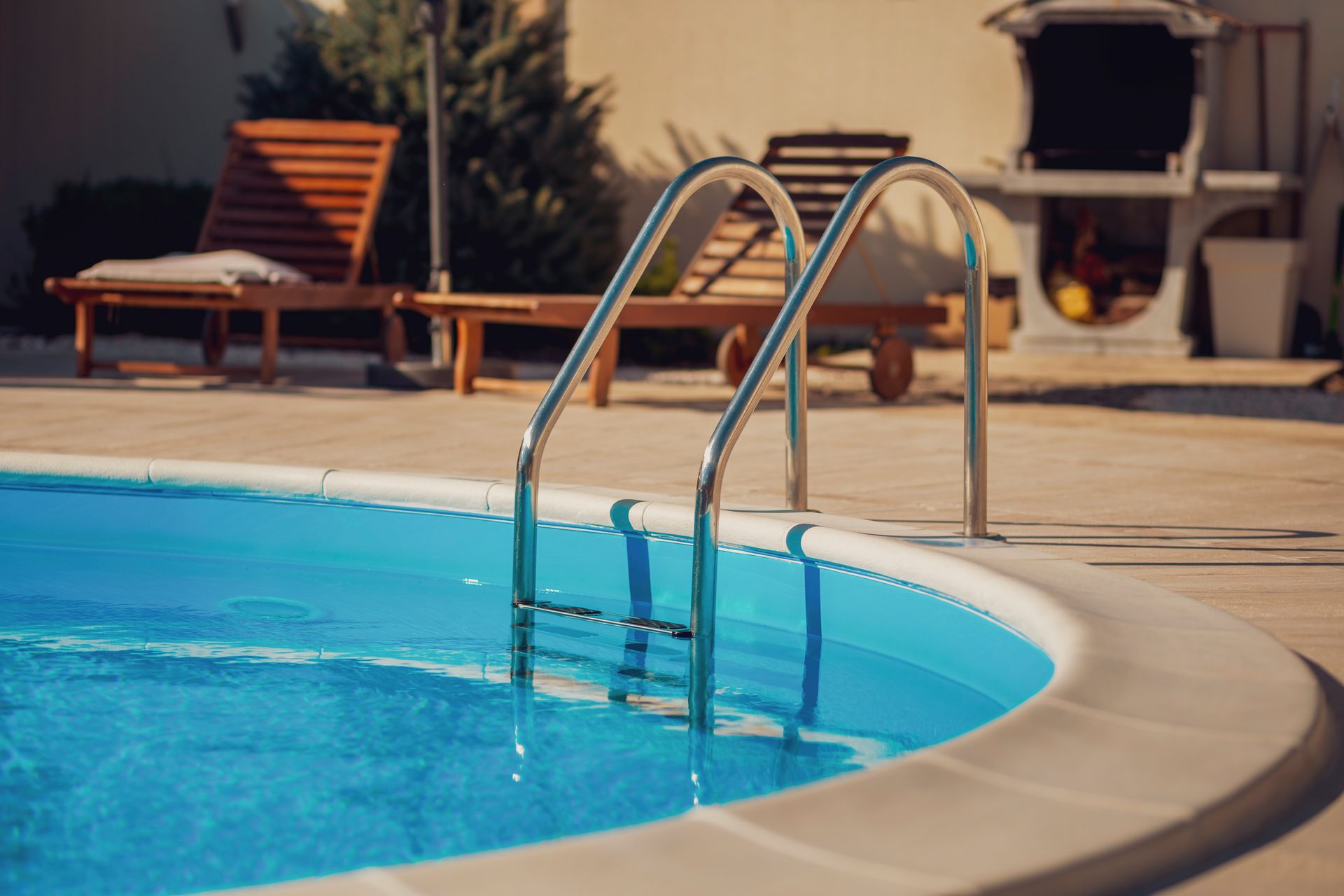 Pool with chrome ladder; wooden lounge chairs and grill in the background on a sunny day.