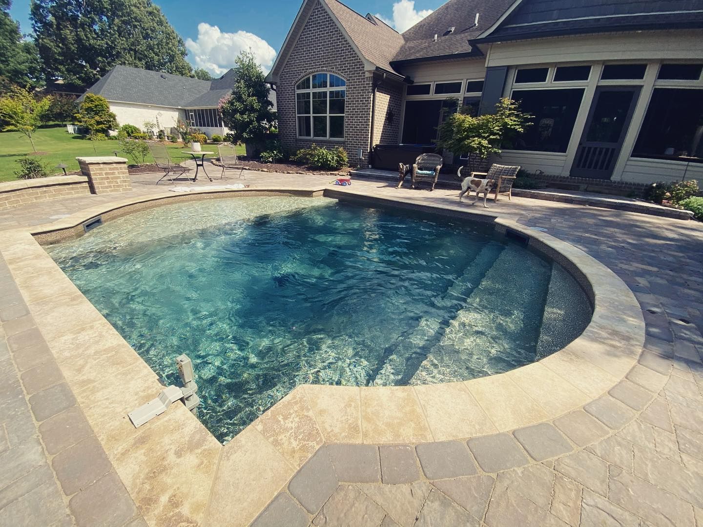 A pool with clear blue water, stone patio, and steps, set in a backyard with a house and sunny sky.