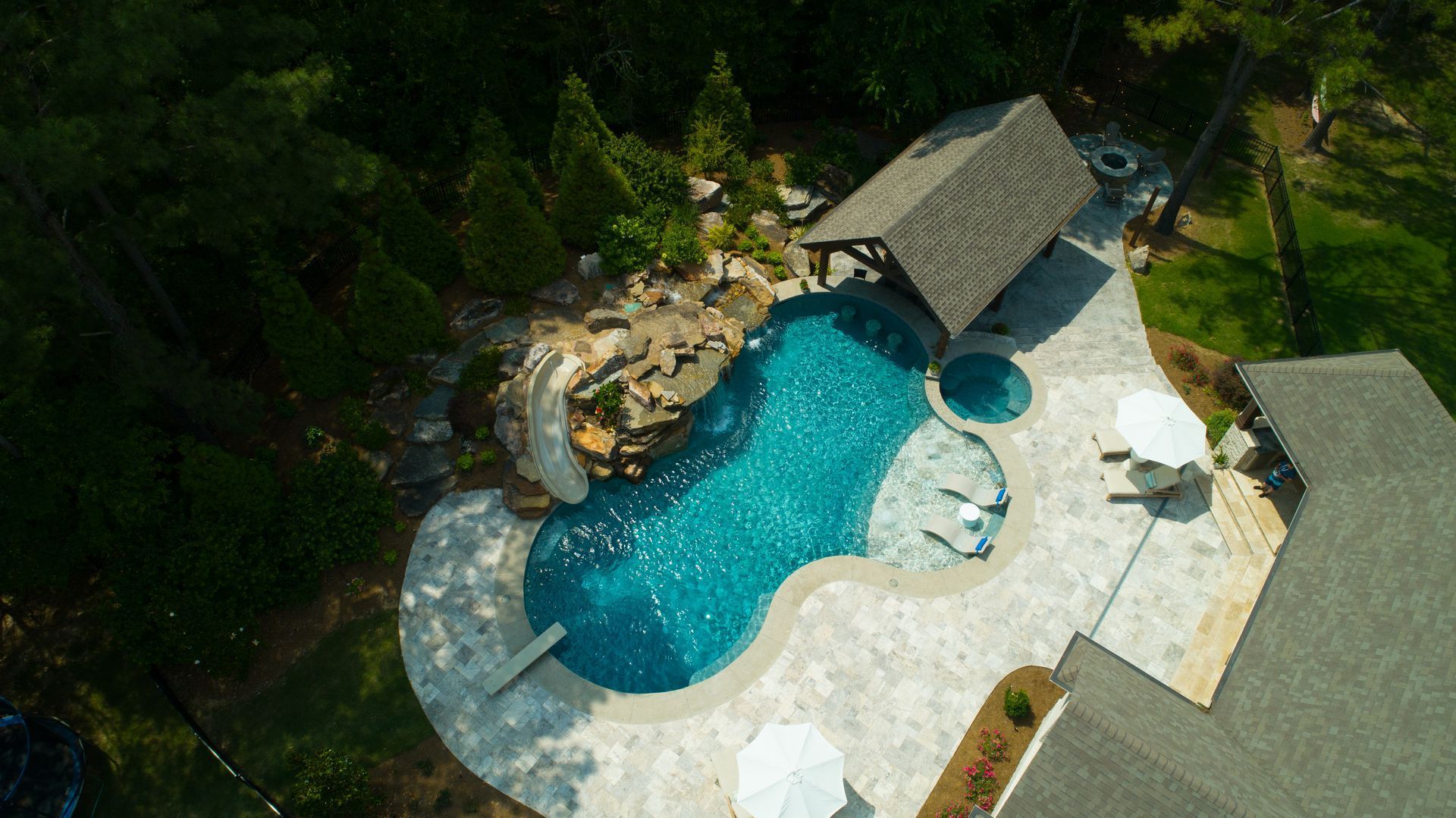 Aerial view of a luxurious pool with a rock waterfall, cabana, and patio area surrounded by trees.